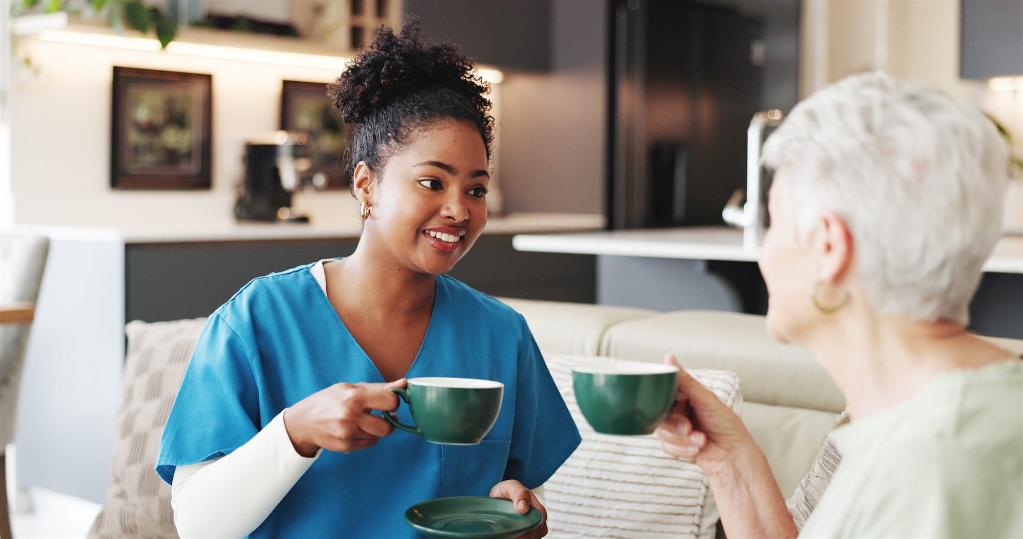A caregiver in scrubs sharing a warm moment over tea with an older adult