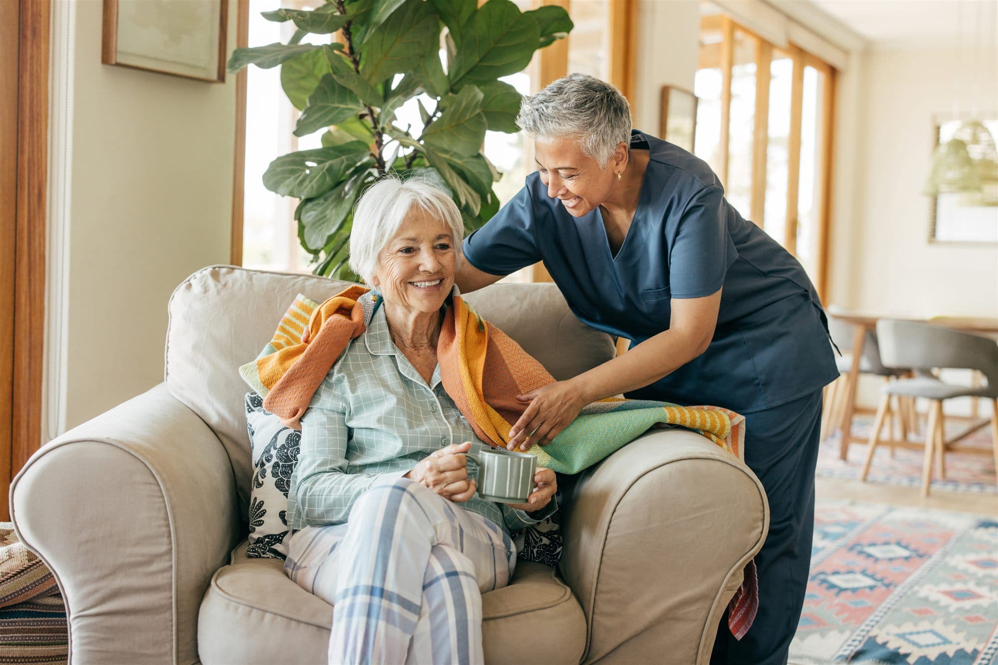 A caregiver wrapping a blanket around an older adult relaxing at home